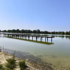 Drewniane molo w Krasnosielcu w letni, słoneczny dzień. Woda spokojna, odbija niebo i drzewa na horyzoncie.