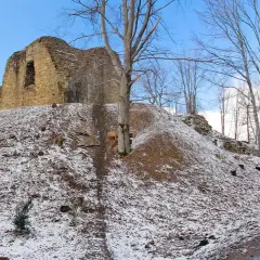 Ruiny zamku Lanckorona na wzgórzu pokrytym śniegiem. Kamienne mury wznoszą się na tle błękitnego nieba, a wokół rosną nagie drzewa.
