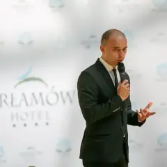 Mężczyzna w garniturze przemawia na tle logo Hotel Arłamów. Właściciel hotelu z pasją opowiada o swoich planach.
