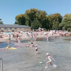 Słoneczny dzień na powidz plaży. Ludzie opalają się, kąpią i bawią w wodzie. W tle budynek i drzewa.