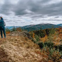 Para turystów na szczycie, podziwiająca jesienny krajobraz gór. Na pierwszym planie kamień i trawa, w tle lasy w barwach jesieni.