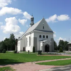 Kościół w Dulczy Wielkiej, z wieżą i krzyżem, na tle błękitnego nieba z chmurami. Obok tablica z wizerunkiem młodego mężczyzny.
