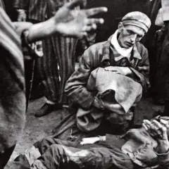 W obozie Majdanek jeden więzień klęczy nad drugim, który leży na ziemi. W tle widać innych więźniów w pasiakach.