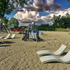 Pusta lubczyna plaża z placem zabaw i leżakami. Kobieta siedzi na ławce, patrząc na telefon.