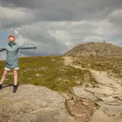 Kobieta w pomarańczowych rękawiczkach i czerwonej opasce cieszy się wolnością na szczycie góry. W oddali widać kamienisty szczyt z krzyżem, a ścieżka prowadzi przez trawiaste zbocza. To idealne miejsce dla krowiarki, która szuka spokoju.