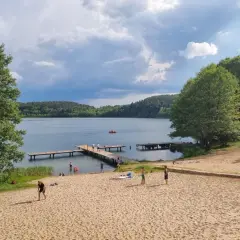 Plaża nad jeziorem z pomostami, drzewami i ludźmi odpoczywającymi na piasku.