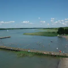 Wypływa z jeziora roś, a plażowicze cieszą się słońcem i wodą. Widać pomosty, łodzie i zielone brzegi.