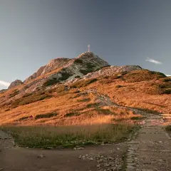 Kamienisty szlak na Giewont w złotym świetle zachodzącego słońca. Widoczny krzyż na szczycie i ławki dla turystów.