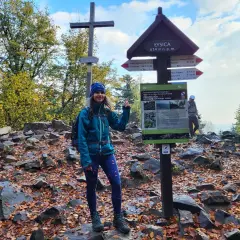 Kobieta na szczycie Łysica, 614 m n.p.m., wśród kamieni i jesiennych liści. Widoczny drogowskaz i krzyż.
