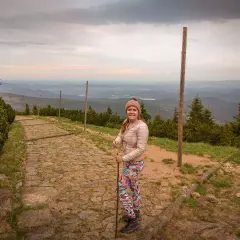 Uśmiechnięta turystka z kijkami na kamienistej ścieżce, podziwia widok na góry. Widać, ile się wchodzi na Śnieżkę.