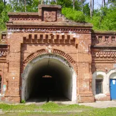Wejście do tunelu w twierdzy Osowiec. Czerwona cegła, łukowate przejście, zieleń na dachu.