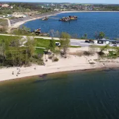 Słoneczny dzień nad zalewem Mietkowskim. Ludzie spacerują po piaszczystej plaży, a w oddali widać budynki i sprzęt pływający.