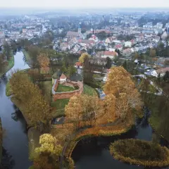 Zamek międzyrzecz otoczony jesiennymi drzewami i wodą, z miastem w tle.