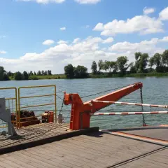 Prom na rzece, z widokiem na zielony brzeg i chmury. W oddali widać drzewa, a na promie pomarańczowy dźwig i znak STOP. To widok z okolic Piotrkowa Kujawskiego.