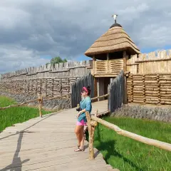 Kobieta na drewnianej kładce podziwia zrekonstruowaną osadę Biskupin. W oddali widać wały obronne i chaty.