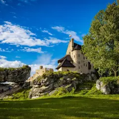 Kamienny zamek na skale, z polską flagą, otoczony zielenią i skalnymi formacjami. W oddali widać łuk skalny, przez który przechodzą turyści. To malownicze mirów bobolice.