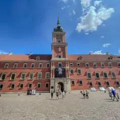 Zamek Królewski w Warszawie, z czerwoną fasadą i wieżą zegarową, na tle błękitnego nieba z chmurami. Plac przed zamkiem tętni życiem.