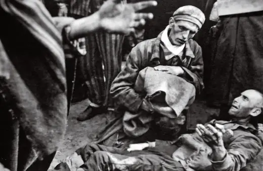 W obozie Majdanek jeden więzień klęczy nad drugim, który leży na ziemi. W tle widać innych więźniów w pasiakach.