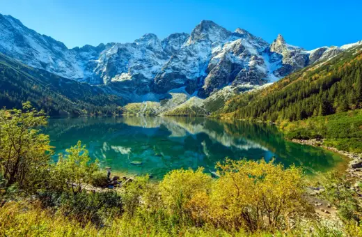 Morskie Oko, Tatry. Turkusowa tafla jeziora odbija ośnieżone szczyty. Widoczna głębokość wody i jesienna roślinność.