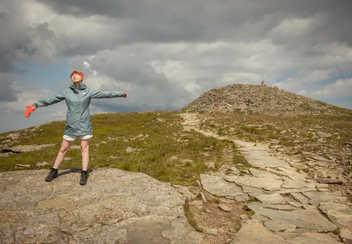 Kobieta w pomarańczowych rękawiczkach i czerwonej opasce cieszy się wolnością na szczycie góry. W oddali widać kamienisty szczyt z krzyżem, a ścieżka prowadzi przez trawiaste zbocza. To idealne miejsce dla krowiarki, która szuka spokoju.