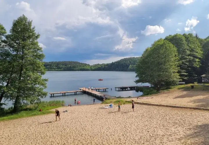 Plaża nad jeziorem z pomostami, drzewami i ludźmi odpoczywającymi na piasku.