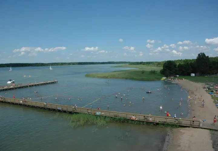 Wypływa z jeziora roś, a plażowicze cieszą się słońcem i wodą. Widać pomosty, łodzie i zielone brzegi.