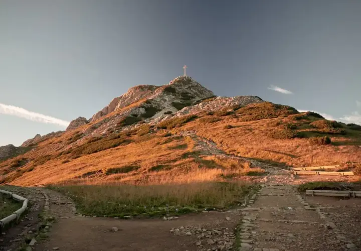 Kamienisty szlak na Giewont w złotym świetle zachodzącego słońca. Widoczny krzyż na szczycie i ławki dla turystów.