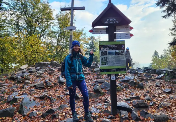 Kobieta na szczycie Łysica, 614 m n.p.m., wśród kamieni i jesiennych liści. Widoczny drogowskaz i krzyż.