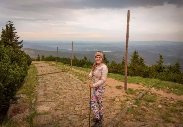 Uśmiechnięta turystka z kijkami na kamienistej ścieżce, podziwia widok na góry. Widać, ile się wchodzi na Śnieżkę.