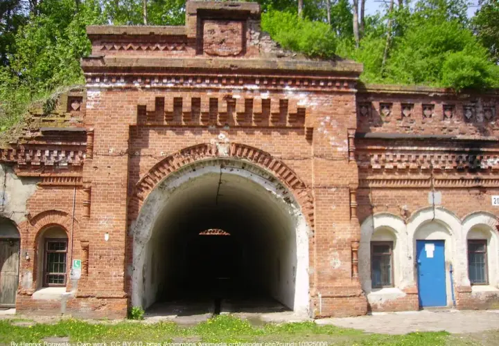 Wejście do tunelu w twierdzy Osowiec. Czerwona cegła, łukowate przejście, zieleń na dachu.