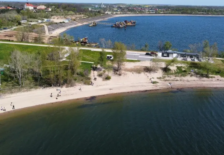 Słoneczny dzień nad zalewem Mietkowskim. Ludzie spacerują po piaszczystej plaży, a w oddali widać budynki i sprzęt pływający.