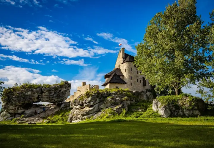 Kamienny zamek na skale, z polską flagą, otoczony zielenią i skalnymi formacjami. W oddali widać łuk skalny, przez który przechodzą turyści. To malownicze mirów bobolice.