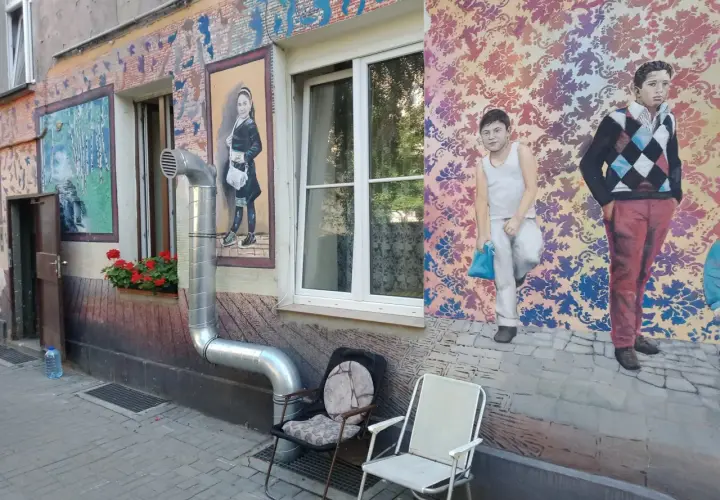 Kolorowe murale z postaciami dzieci i wzorami zdobią kamienicę na Nadodrzu. Dwa krzesła stoją przed ścianą.