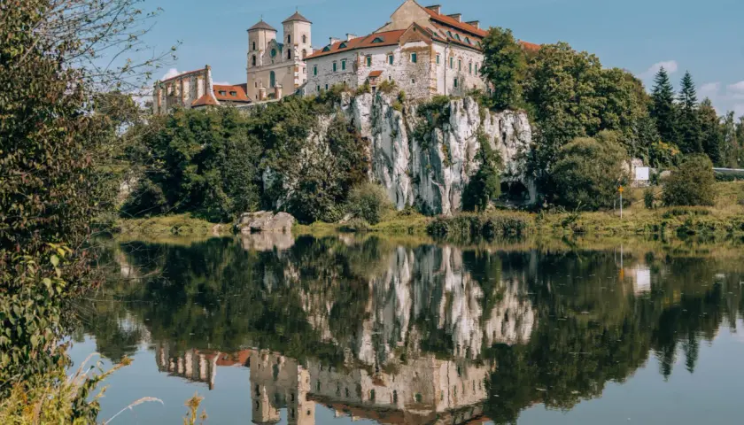 Majestatyczne opactwo na skale odbija się w spokojnej tafli rzeki, otoczone zielenią drzew.