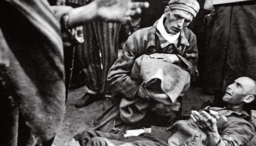 W obozie Majdanek jeden więzień klęczy nad drugim, który leży na ziemi. W tle widać innych więźniów w pasiakach.
