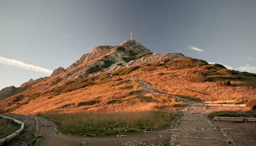 Kamienisty szlak na Giewont w złotym świetle zachodzącego słońca. Widoczny krzyż na szczycie i ławki dla turystów.