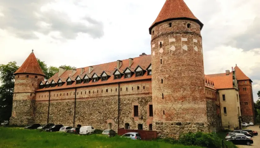 Zamek w Bytowie, potężna ceglana twierdza z charakterystycznymi wieżami i kamiennymi murami, otoczony zielenią i parkingiem.