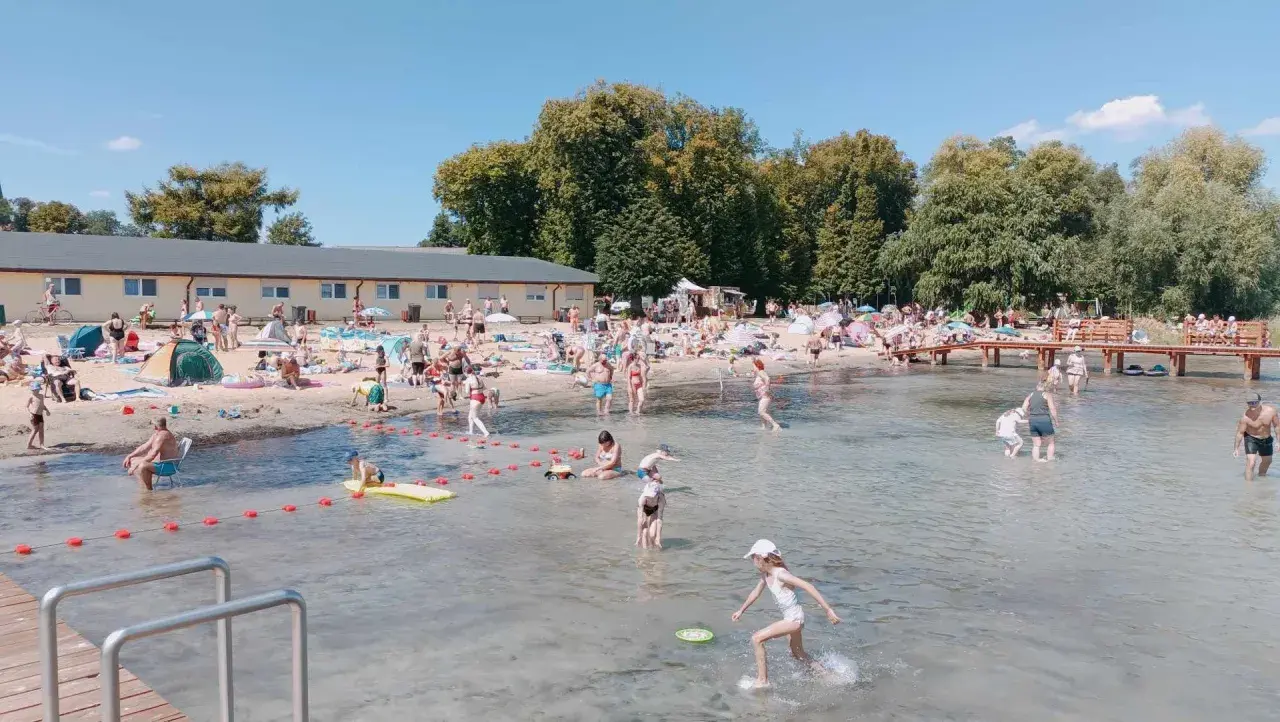 Słoneczny dzień na powidz plaży. Ludzie opalają się, kąpią i bawią w wodzie. W tle budynek i drzewa.