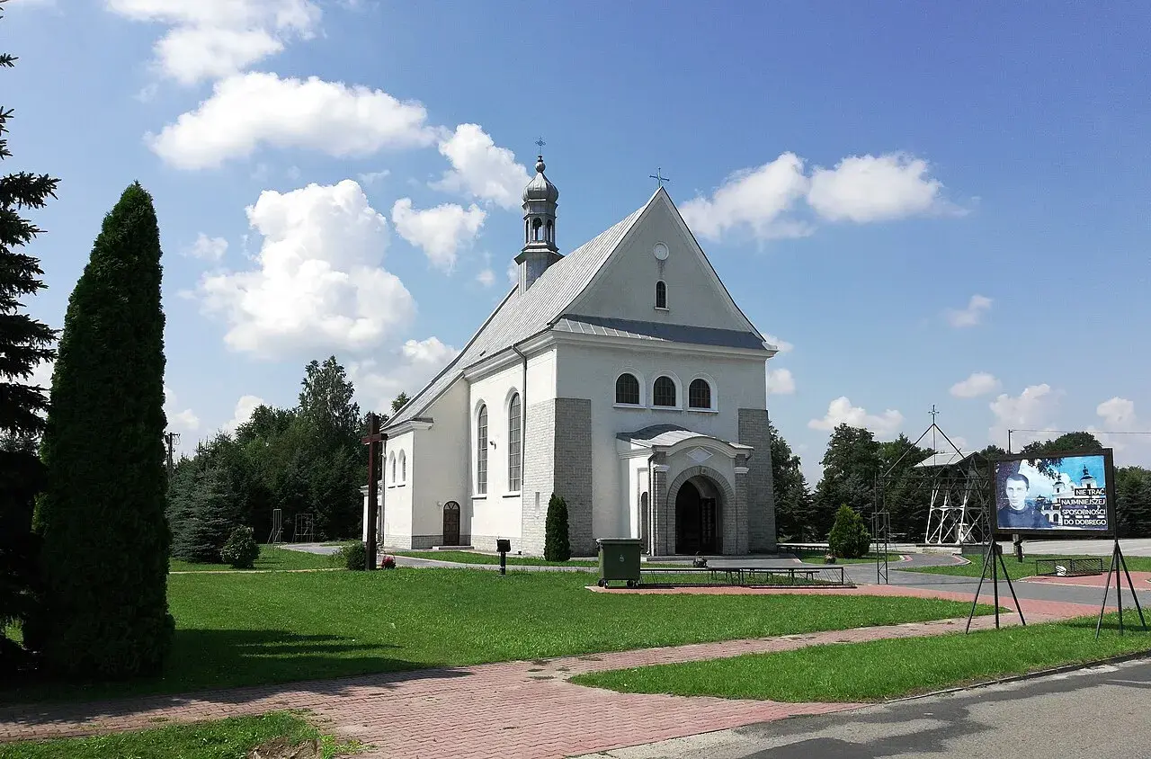 Kościół w Dulczy Wielkiej, z wieżą i krzyżem, na tle błękitnego nieba z chmurami. Obok tablica z wizerunkiem młodego mężczyzny.