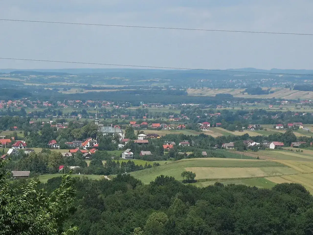 Widok na malowniczą wieś Przytkowice z kościołem w centrum, otoczoną zielonymi wzgórzami i polami.