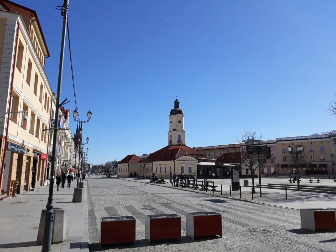 Słoneczny dzień na Rynku Kościuszki w Białymstoku. W oddali wieża ratusza, po bokach kamienice i brukowana nawierzchnia.