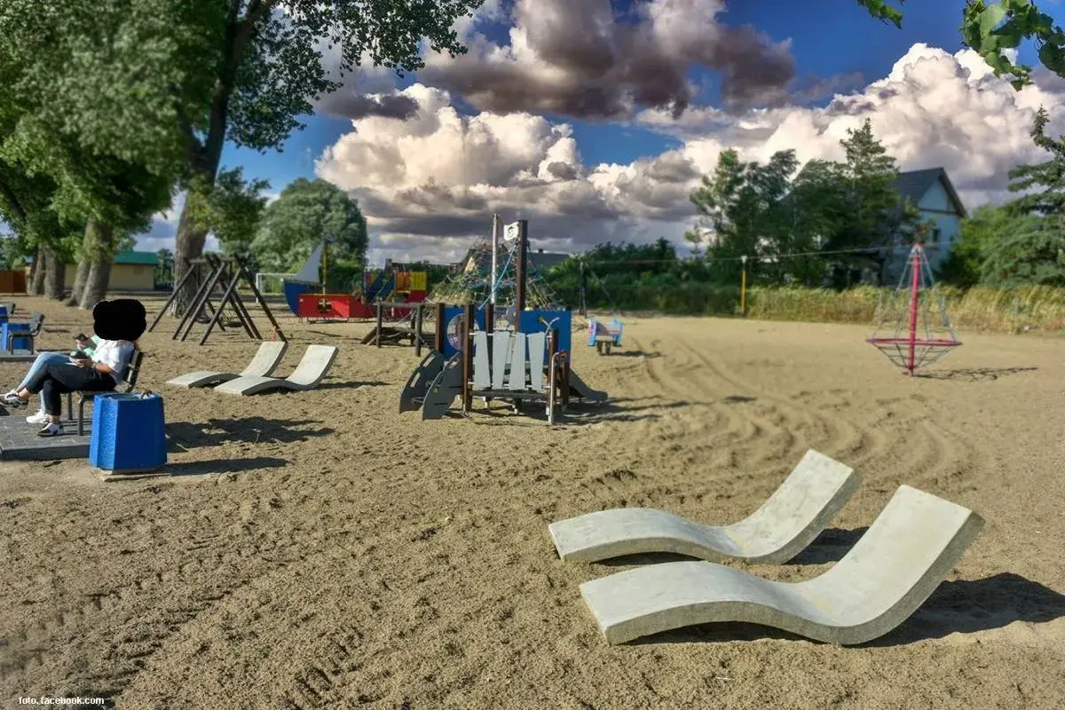 Pusta lubczyna plaża z placem zabaw i leżakami. Kobieta siedzi na ławce, patrząc na telefon.