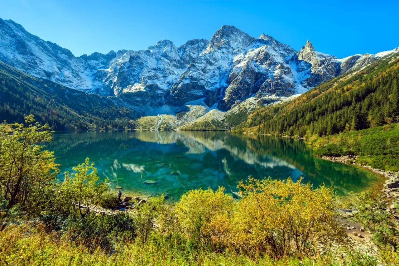 Morskie Oko, Tatry. Turkusowa tafla jeziora odbija ośnieżone szczyty. Widoczna głębokość wody i jesienna roślinność.