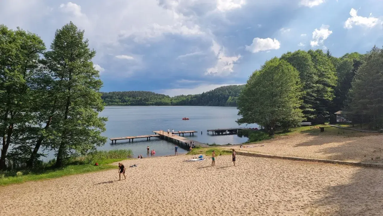 Plaża nad jeziorem z pomostami, drzewami i ludźmi odpoczywającymi na piasku.