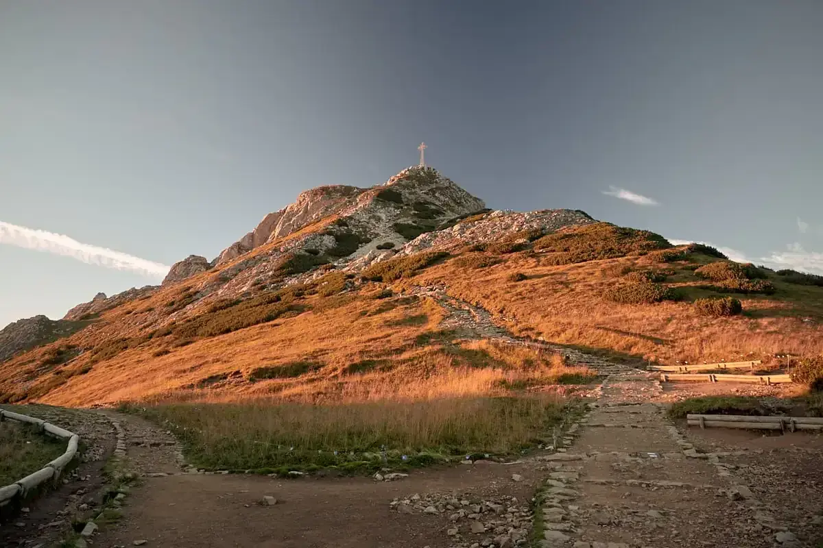 Kamienisty szlak na Giewont w złotym świetle zachodzącego słońca. Widoczny krzyż na szczycie i ławki dla turystów.