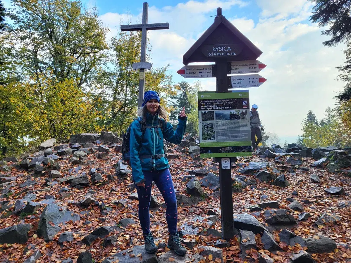 Kobieta na szczycie Łysica, 614 m n.p.m., wśród kamieni i jesiennych liści. Widoczny drogowskaz i krzyż.