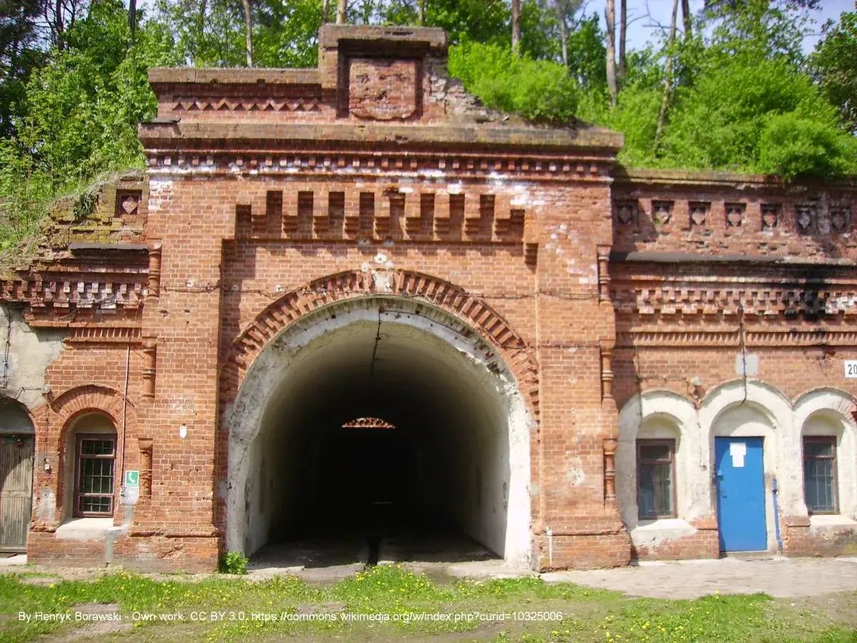 Wejście do tunelu w twierdzy Osowiec. Czerwona cegła, łukowate przejście, zieleń na dachu.