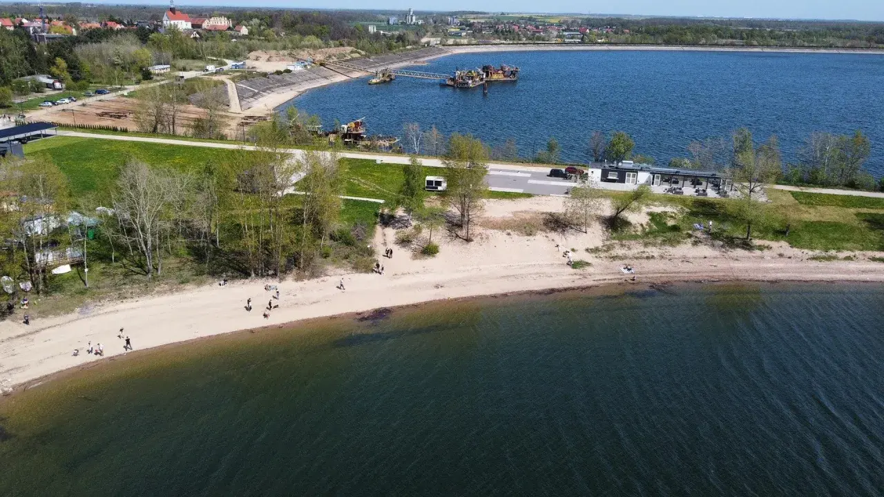 Słoneczny dzień nad zalewem Mietkowskim. Ludzie spacerują po piaszczystej plaży, a w oddali widać budynki i sprzęt pływający.