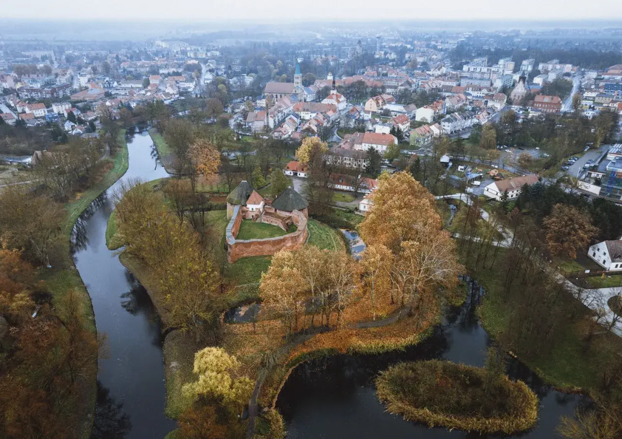 Zamek międzyrzecz otoczony jesiennymi drzewami i wodą, z miastem w tle.