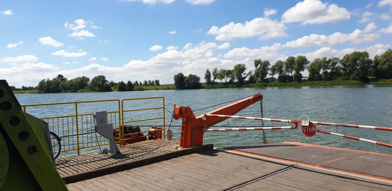 Prom na rzece, z widokiem na zielony brzeg i chmury. W oddali widać drzewa, a na promie pomarańczowy dźwig i znak STOP. To widok z okolic Piotrkowa Kujawskiego.